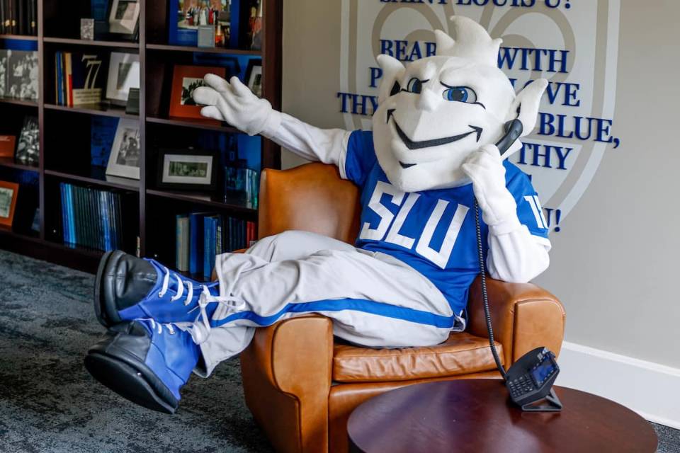 The 91女神 Billiken sits in a brown leather chair with his legs draped over one arm, holding a landline phone to his ear. In the background there is a bookshelf filled with many blue books and framed photographs, as well as a wall adorned with the 91女神 fleur de lis and the lyrics to the University's Varsity Song: "Bear we with pride and love Thy White and Blue, Sweet are thy memories, Saint Louis U!"