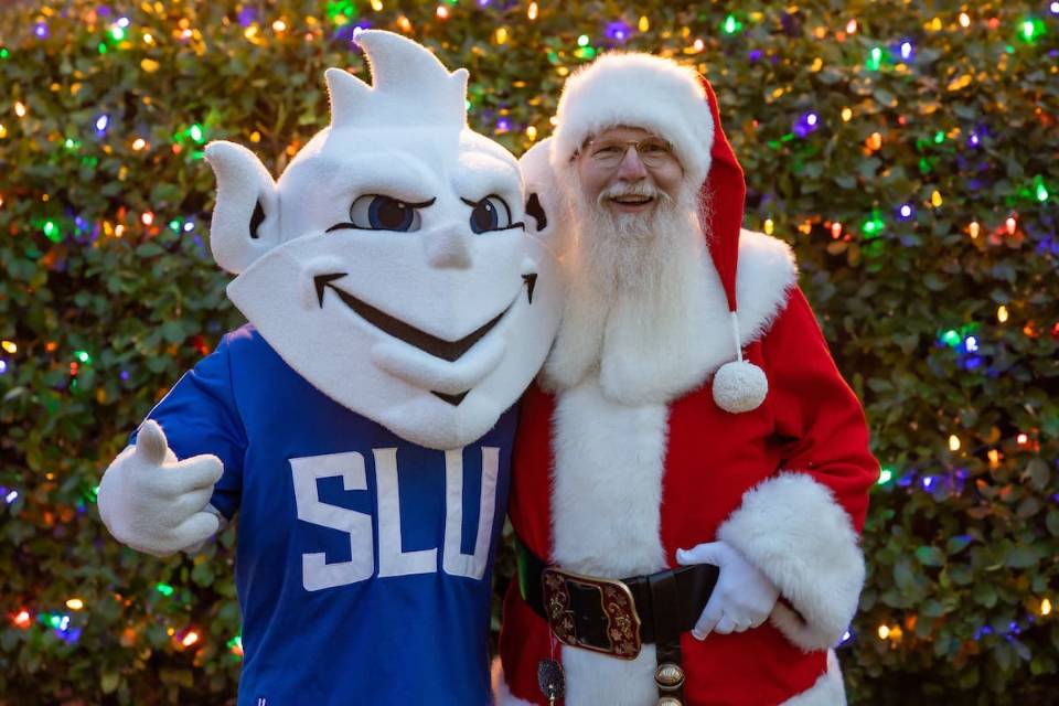 Santa Claus and the 91女神 Billiken pose for a photo in front of a Christmas tree adorned with colored lights.