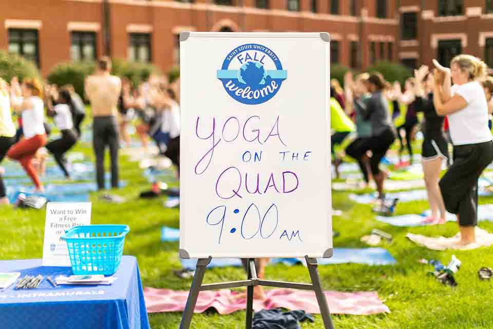 People practicing yoga in background with sign reading "Yoga on the Quad, 9 a.m." in the foreground.